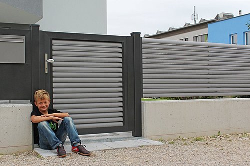 Gehtür aus Lamellen in grau mit Rahmenoptik in anthrazit, Modell Plissée, mit passendem Briefkasten, Kind sitzt vor Zaun am Boden