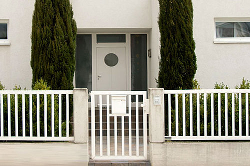Schlichter Einzelstabzaun in weiß mit passender Gartentüre und integriertem Briefkasten, Modell Siena, vor einem weißen, modernen Haus