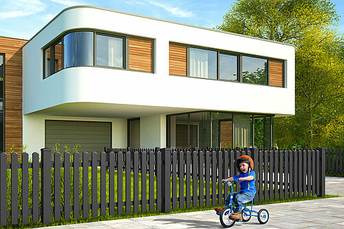 Lattenzaun aus Aluminium in anthrazit mit Rundkappen, Modell Merlin von GUARDI, vor einem modernen, weißen Haus, Kind fährt vor Zaun mit Dreirad