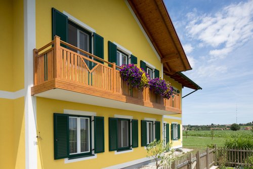 Balkongeländer in Holzoptik im Modell Kitzbühel, der Balkon ist auf einem gelben Haus montiert, die Fenster haben grüne Rahmen