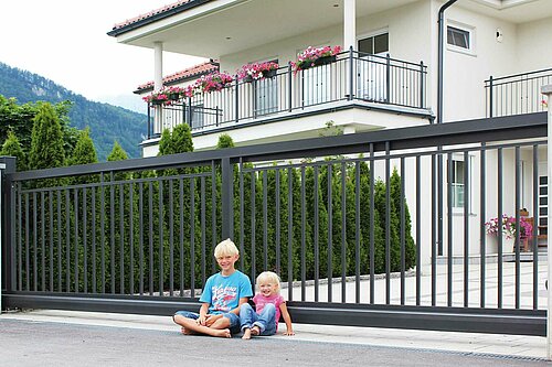  elektrisches Schiebetor aus Einzelstäben in anthrazit, Modell Toskana von Guardi, vor weißem Haus mit großem Balkon, zwei Kinder sitzen vor dem Tor