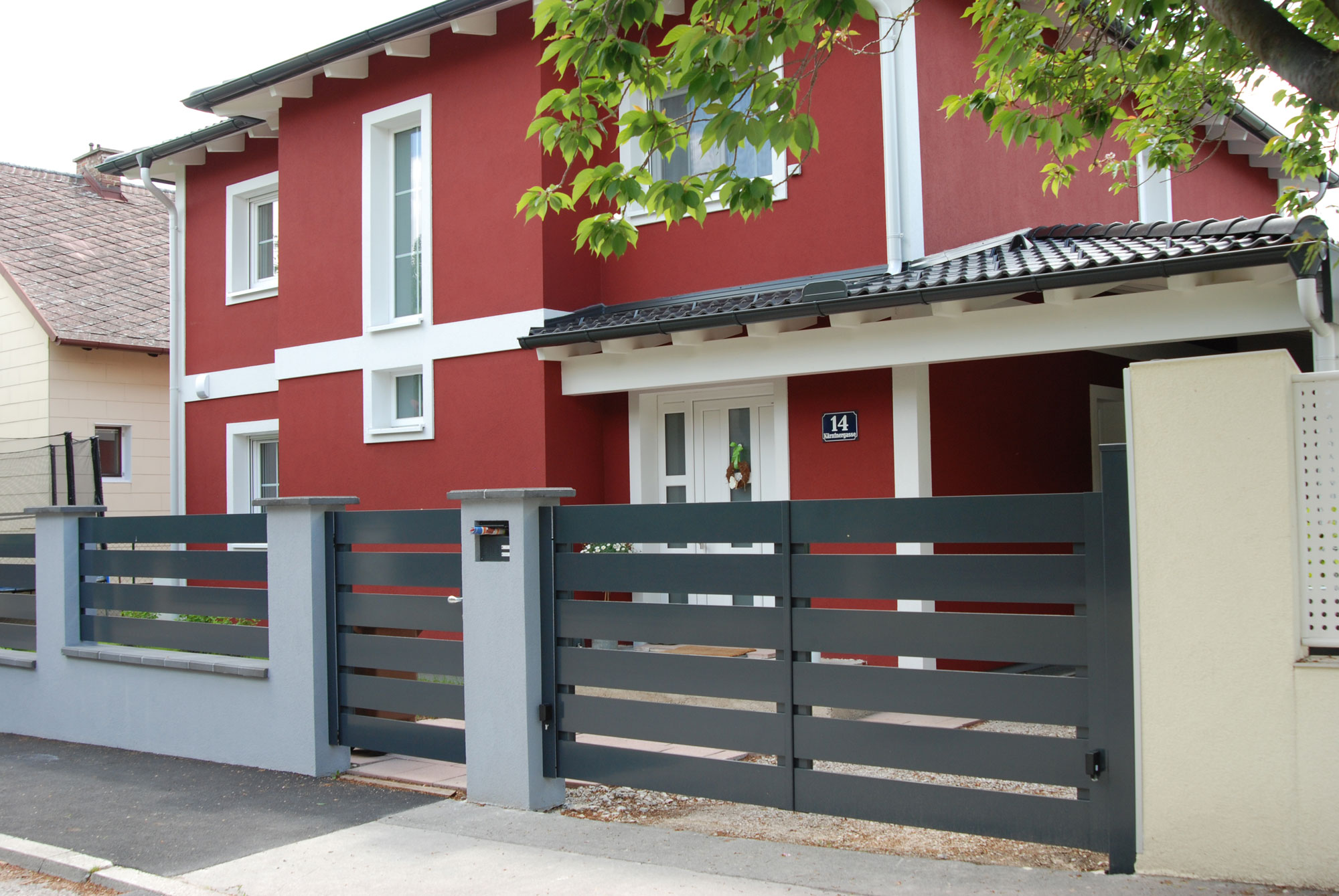 Moderner Gartenzaun mit Flügeltor und Gehtüre mit Querlatten aus Aluminium begrenzt ein Grundstück vor einem roten Haus